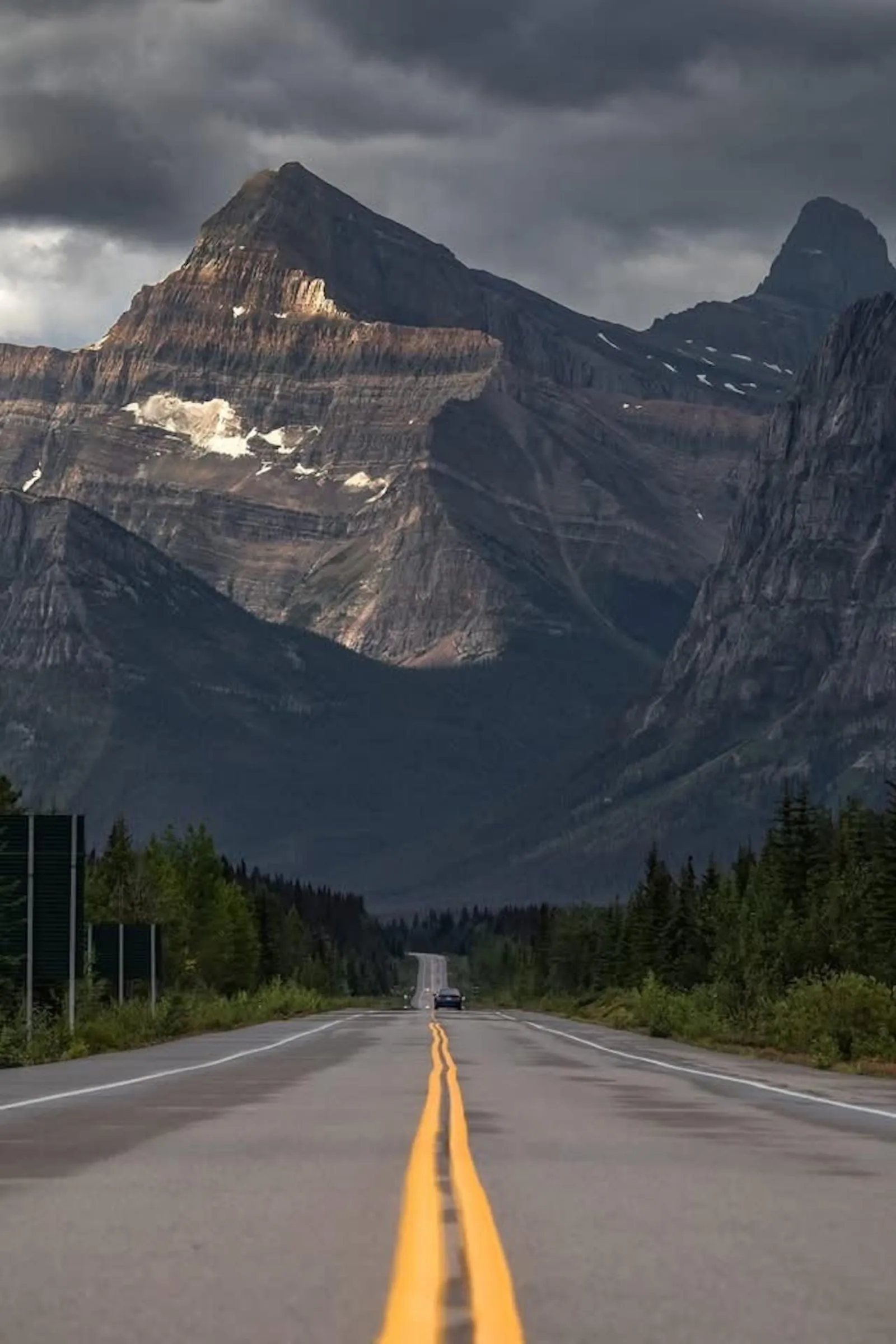 The Icefields Parkway running straight toward layered Rockies peaks under heavy cloud