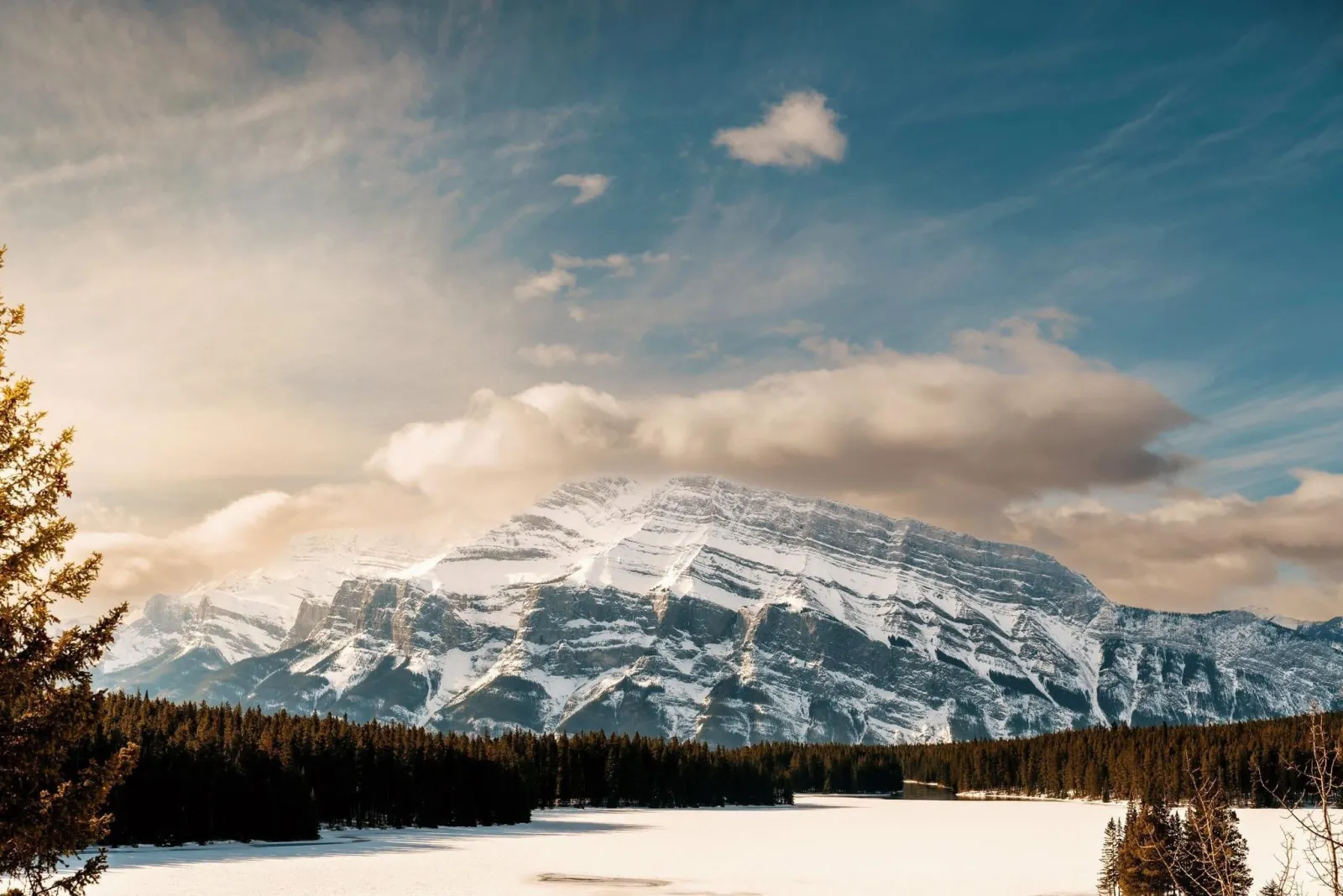 A snow-covered Rockies massif above a frozen lake and dark spruce forest in winter light