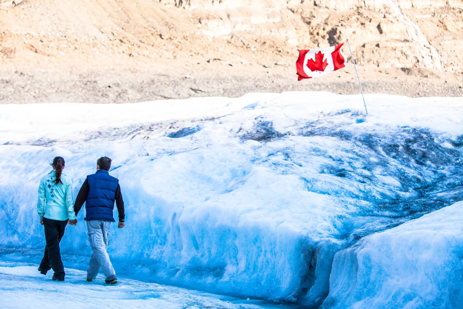 Two visitors walking on blue glacier ice beside the Canadian flag marking the roped visitor area