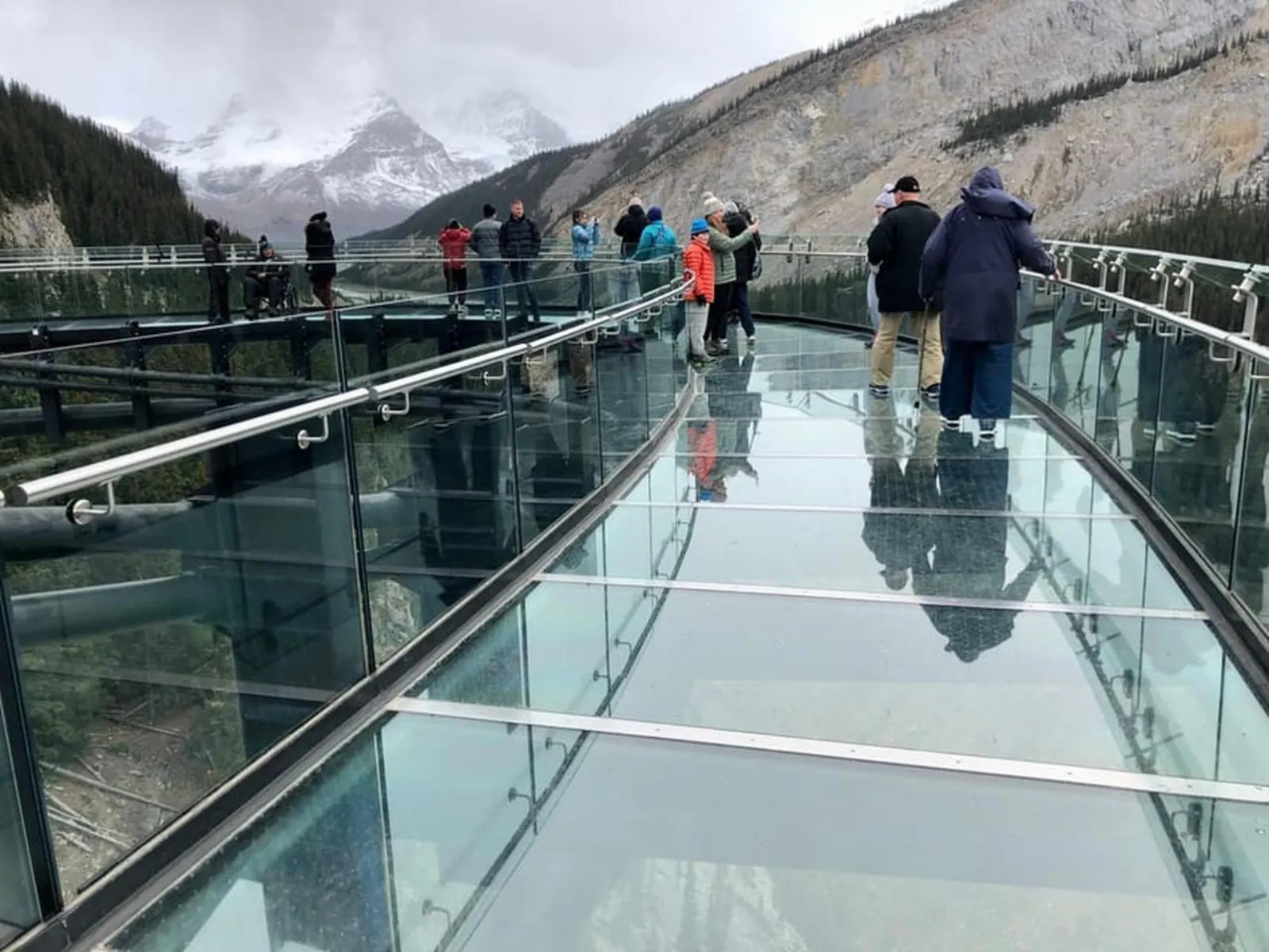 Visitors on the Skywalk's glass deck on an overcast day, snow-dusted peaks in the distance