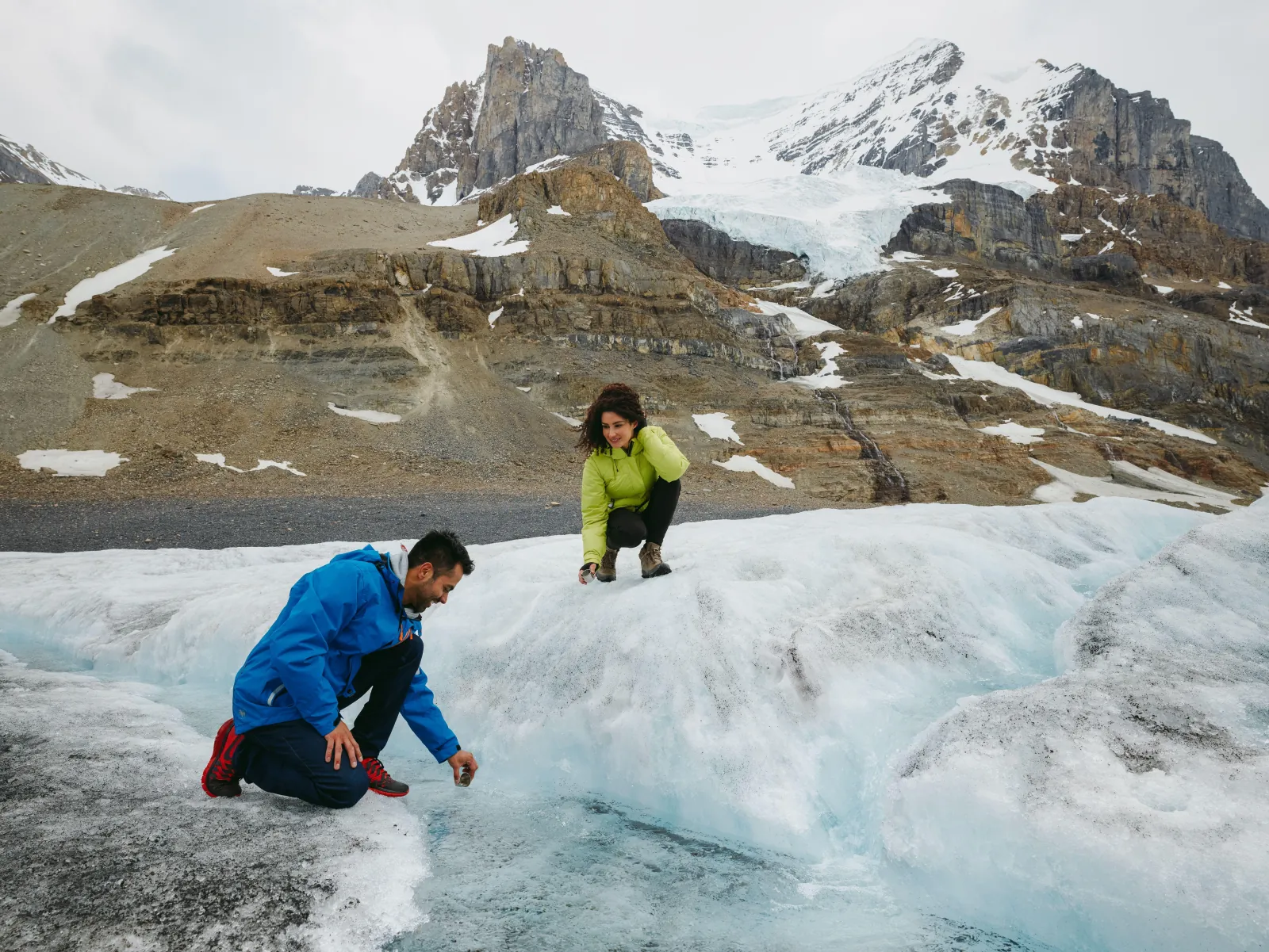 Two visitors crouching to drink meltwater running across the Athabasca Glacier